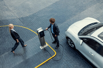 Aerial view progressive lifestyle of businesspeople with electric car connected to charging station at city center public parking car. Eco friendly rechargeable car powered by alternative clean energy