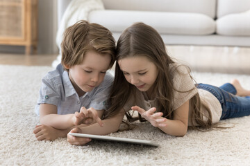 Wall Mural - Curious little brother and sister kids using tablet for Internet communication, lying on warm soft carpeted floor, playing video game, browsing blogs, chatting, enjoying leisure alone