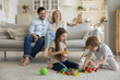 © fizkes - Two cheerful excited sibling kids playing with toy blocks, colorful cubes on soft carpeted floor, constructing towers, laughing on heap of plastic pieces. Couple of parents watching little children