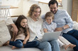 © fizkes - Excited parents and two happy little kids using laptop at home for Internet communication, wireless online connection, relaxing on sofa together, watching movie, smiling, laughing