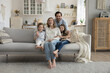 © fizkes - Joyful family couple and little son and daughter kids sitting on sofa, resting on comfortable couch at cozy home, hugging with love, joy, looking at camera, smiling. Full length wide shot