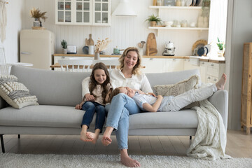 Positive young mom enjoying motherhood, leisure time at home with sweet little son and daughter, resting on soft couch with children, talking to kids, smiling, laughing