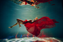 Art Work. A Slender, Tanned Girl With An Athletic Figure And Blond Hair, With Red Material And Light Underwear, Dives And Spins Underwater In The Pool. Aesthetic Image For Your Design Or Decoration.