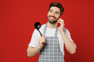 Sticker - Young male housewife housekeeper chef cook baker man wear grey apron talk speak on mobile cell phone hold soup black ladle dipper isolated on plain red color background studio. Cooking food concept.