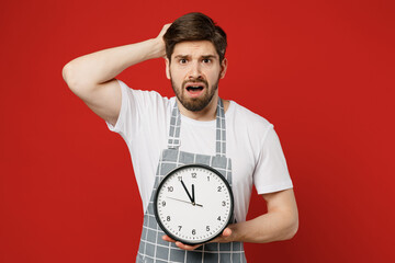 Sticker - Young confused scared shocked displeased disappointed male housewife housekeeper chef cook baker man wear grey apron hold head show clock isolated on plain red background studio. Cooking food concept.