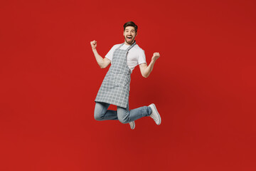 Sticker - Full body overjoyed happy fun cool young male housewife housekeeper chef cook baker man wearing grey apron jumps high do winner gesture isolated on plain red background studio. Cooking food concept.