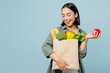 Young woman wear casual clothes hold apple look at brown paper bag with food products after shopping isolated on plain blue cyan background studio portrait. Delivery service from shop or restaurant.