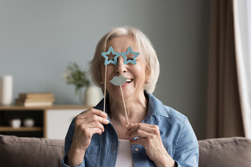 Cheerful happy senior woman applying fake paper moustache and goggles on stick to face, looking at camera, having fun, smiling, laughing, using prop for photo shooting