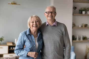 Cheerful positive senior married couple posing at home, standing close, looking at camera, smiling, hugging with love, affection. Indoor portrait of happy elderly husband and wife enjoying friendship