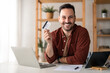 © Dorde - Confident businessman holding in hand showing credit card while sitting in home office. Caucasian businessman advertising banking services, loan or deposits, cashback for purchases from home concept.