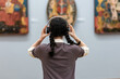 Back view of young woman wearing headphones and contemplates ancient arts. Student visiting gallery or museum. Concept of modern education and culture