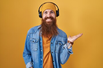 Wall Mural - Caucasian man with long beard listening to music using headphones smiling cheerful presenting and pointing with palm of hand looking at the camera.