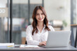 © amnaj - Happy Asian businesswoman sitting in office with her laptop computer working attentively with various documents on her desk.
