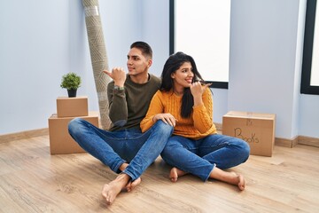 Poster - Young couple sitting on the floor at new home smiling with happy face looking and pointing to the side with thumb up.