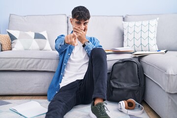 Sticker - Young hispanic man sitting on the floor studying for university laughing at you, pointing finger to the camera with hand over mouth, shame expression