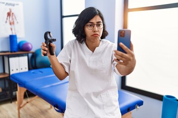 Wall Mural - Young hispanic physiotherapist woman holding hand grip to train muscle doing video call depressed and worry for distress, crying angry and afraid. sad expression.