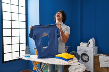 Poster - Hispanic man with curly hair ironing holding burned iron shirt at laundry room angry and mad screaming frustrated and furious, shouting with anger looking up.