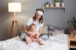 © sementsova321 - Photo of joyful cheerful mother and child laughing and playing together while sitting on bed, woman wearing pajama and sleeping mask having fun with her infant kid.