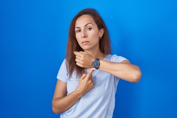 Poster - Brunette woman standing over blue background in hurry pointing to watch time, impatience, looking at the camera with relaxed expression