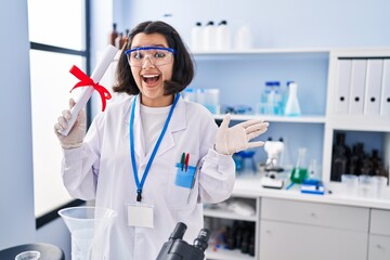 Canvas Print - Young hispanic woman working at scientist laboratory holding degree celebrating achievement with happy smile and winner expression with raised hand