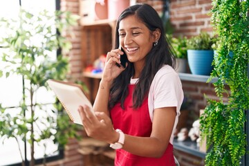 Poster - Young beautiful hispanic woman florist talking on smartphone reading notebook at flower shop