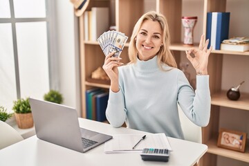 Poster - Young caucasian woman using laptop holding dollars banknotes doing ok sign with fingers, smiling friendly gesturing excellent symbol