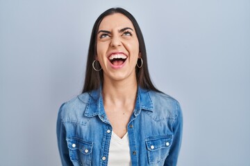 Sticker - Hispanic woman standing over blue background angry and mad screaming frustrated and furious, shouting with anger. rage and aggressive concept.