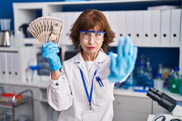 Wall Mural - Middle age woman working at scientist laboratory holding money with open hand doing stop sign with serious and confident expression, defense gesture