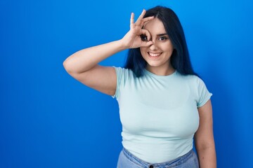 Wall Mural - Young modern girl with blue hair standing over blue background doing ok gesture with hand smiling, eye looking through fingers with happy face.