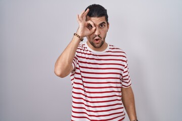 Wall Mural - Hispanic man with long hair standing over isolated background doing ok gesture shocked with surprised face, eye looking through fingers. unbelieving expression.