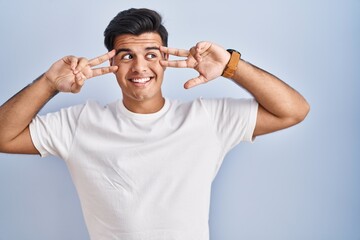 Canvas Print - Hispanic man standing over blue background doing peace symbol with fingers over face, smiling cheerful showing victory