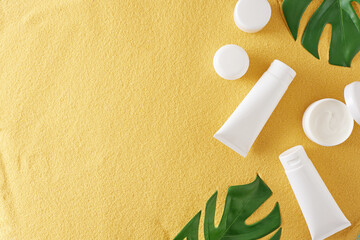 Wall Mural - Sunscreen cosmetic concept. Flat lay photo of cosmetic bottles without label, cream jars and green tropical leaves on sandy background with empty space. Skin care products mockup idea.