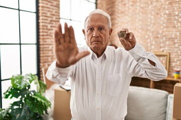 Poster - Senior man with grey hair holding virtual currency bitcoin with open hand doing stop sign with serious and confident expression, defense gesture