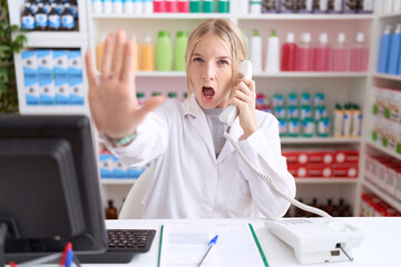 Sticker - Young caucasian woman working at pharmacy drugstore speaking on the telephone doing stop gesture with hands palms, angry and frustration expression