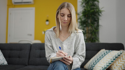 Canvas Print - Young blonde woman writing on notebook sitting on sofa at home