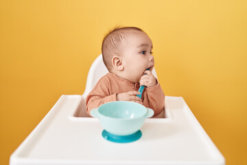 Poster - Adorable hispanic toddler sitting on highchair eating over isolated yellow background