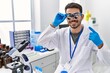 © Krakenimages.com - Young hispanic man working at scientist laboratory wearing magnifying glasses smiling happy pointing with hand and finger