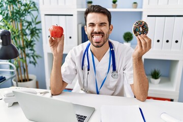 Sticker - Young hispanic dietitian man holding doughnut and apple sticking tongue out happy with funny expression.