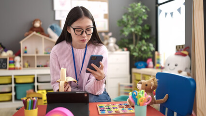 Sticker - Young chinese woman preschool teacher eating banana using smartphone at kindergarten