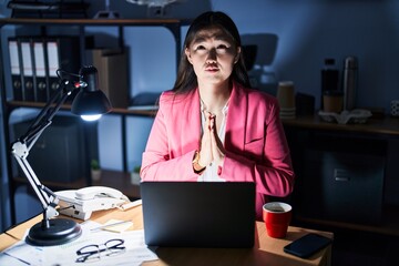 Wall Mural - Chinese young woman working at the office at night begging and praying with hands together with hope expression on face very emotional and worried. begging.
