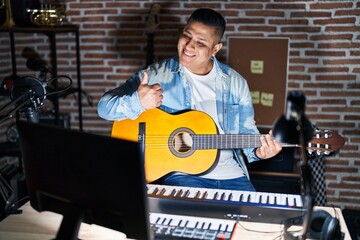 Poster - Hispanic young man playing classic guitar at music studio doing happy thumbs up gesture with hand. approving expression looking at the camera showing success.