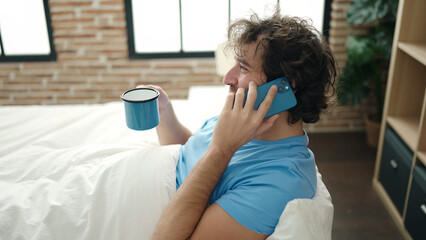 Wall Mural - Young hispanic man talking on smartphone drinking coffee at bedroom