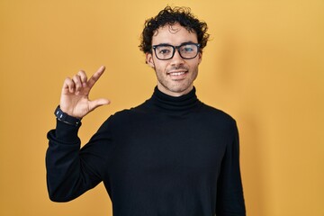 Sticker - Hispanic man standing over yellow background smiling and confident gesturing with hand doing small size sign with fingers looking and the camera. measure concept.