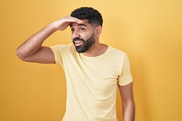 Poster - Hispanic man with beard standing over yellow background very happy and smiling looking far away with hand over head. searching concept.