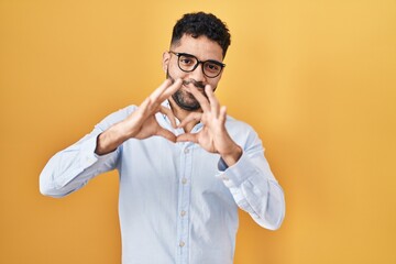 Sticker - Hispanic man with beard standing over yellow background smiling in love doing heart symbol shape with hands. romantic concept.