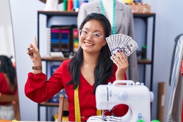 Poster - Asian young woman dressmaker designer holding dollars smiling happy pointing with hand and finger to the side