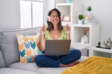 Sticker - Hispanic young woman using laptop at home gesturing finger crossed smiling with hope and eyes closed. luck and superstitious concept.