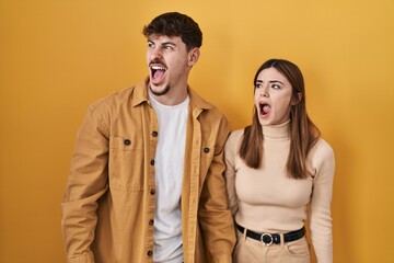 Sticker - Young hispanic couple standing over yellow background angry and mad screaming frustrated and furious, shouting with anger. rage and aggressive concept.