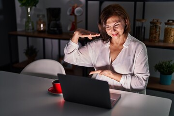 Poster - Middle age hispanic woman using laptop at home at night gesturing with hands showing big and large size sign, measure symbol. smiling looking at the camera. measuring concept.