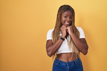 Wall Mural - African american woman with braided hair standing over yellow background laughing nervous and excited with hands on chin looking to the side